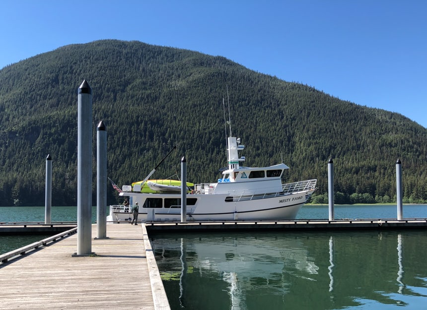 The small ship Misty Fjord floats at a dock, behind her is a forested mountain side and clear blue sky.