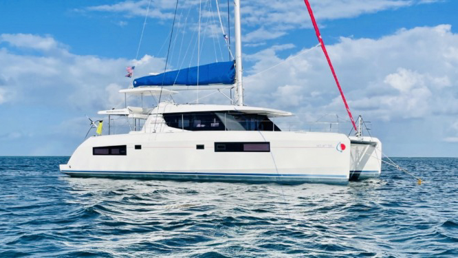 White Belize catamaran Nice Aft Too with blue sail storage jacket & lots of windows sits in calm water on a sunny day.