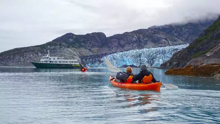 white and green colored small ship anchors near icy teal glacier in Alaska as guests paddle kayaks nearby