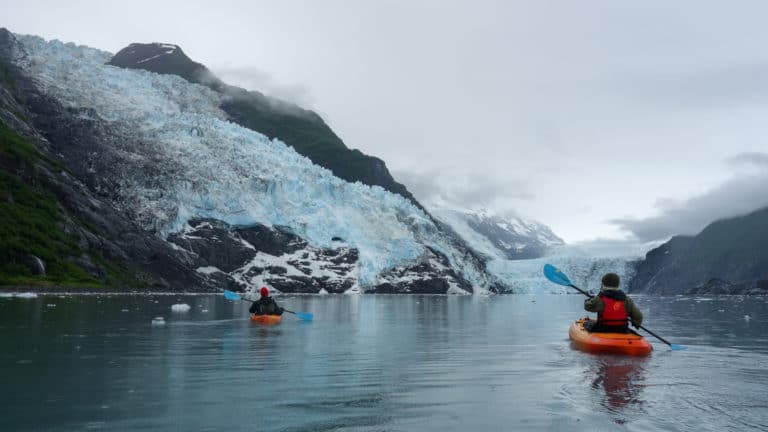 On a Prince William Sound cruise two kayakers paddle towards a massive glacier on an overcast day.