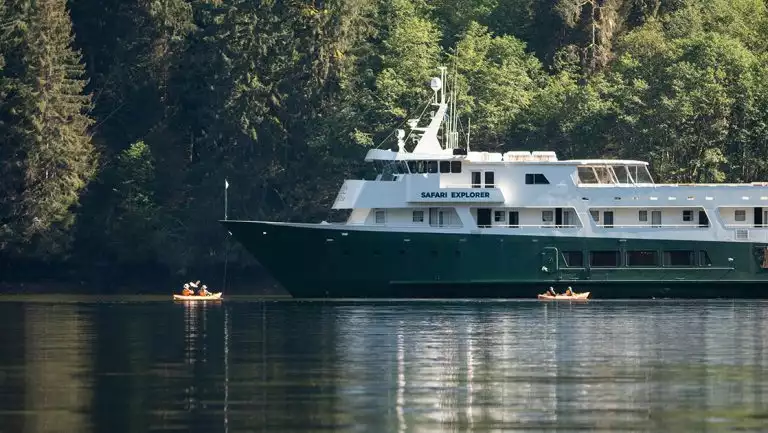 White boat with forest green pained hull Safari Explorer anchors close to a densely forested shoreline in Alaska as guests kayak around