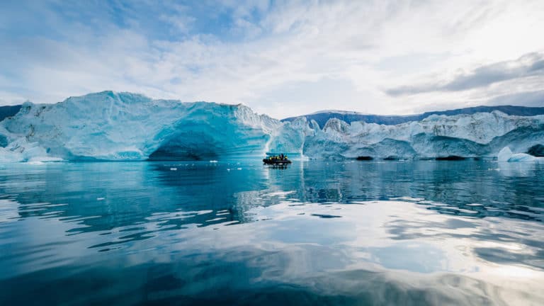 A Zodiac with polar travelers cruises past blue icebergs in a calm bay on a partly sunny day during the South Georgia Antarctic Odyssey Cruise.