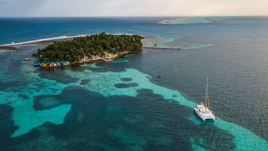 Seen from above a small white Belize catamaran floats in crystal clear ocean water in front of a tiny palm tree covered island, a popular option for private charter.
