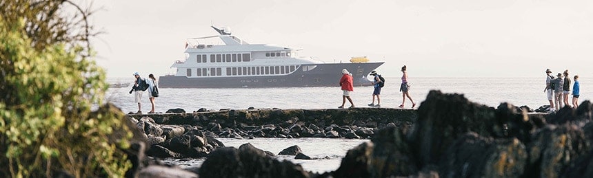 The origin and theory luxury Galapagos yacht floats on the horizon as guests walk the shoreline of an island as part of a daily excursion. 