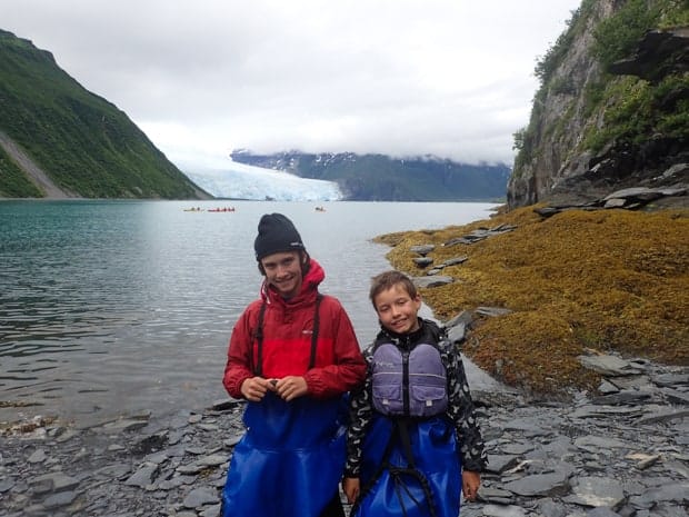Two young boys wearing kayak skirts smiling at the camera in a bay with kayakers and a glacier behind them.