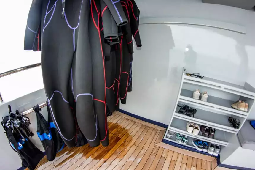 The snorkel station aboard Camila Galapagos trimaran, long black wet suits hang to dry with bags of masks, fins and snorkels, next to them a 6 row shelf with shoes