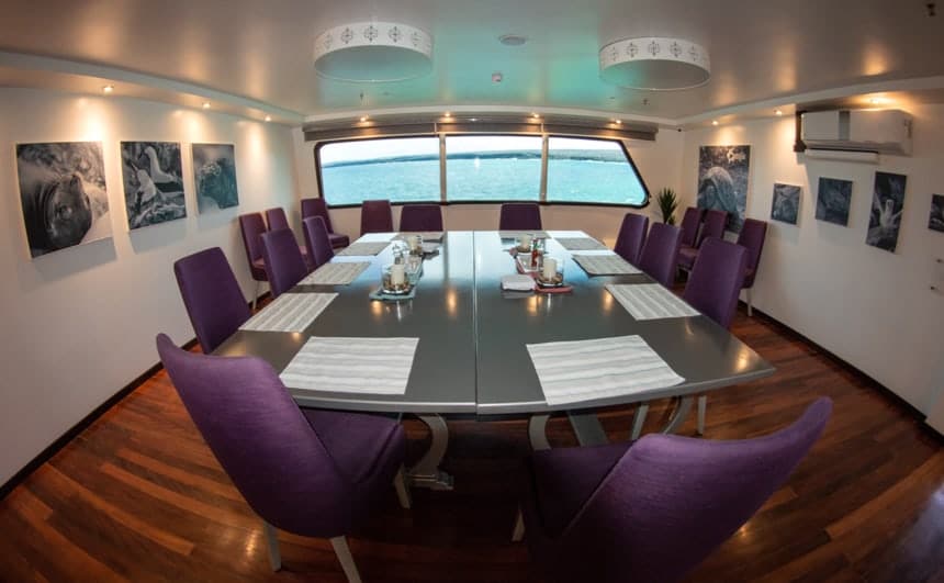 The dining room inside the Camila galapagos trimaran, purple chairs surround a grey table, a set of windows on one wall, and black and white painting are found on the others.