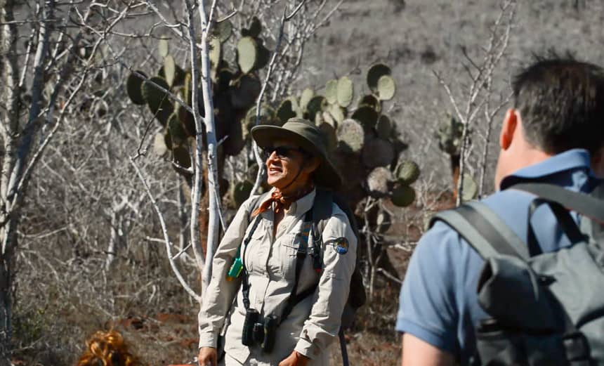 A female galapagos naturalist guide stands and speaks in front of a cruise group on rabida island, behind her are cactus and palo santos trees.