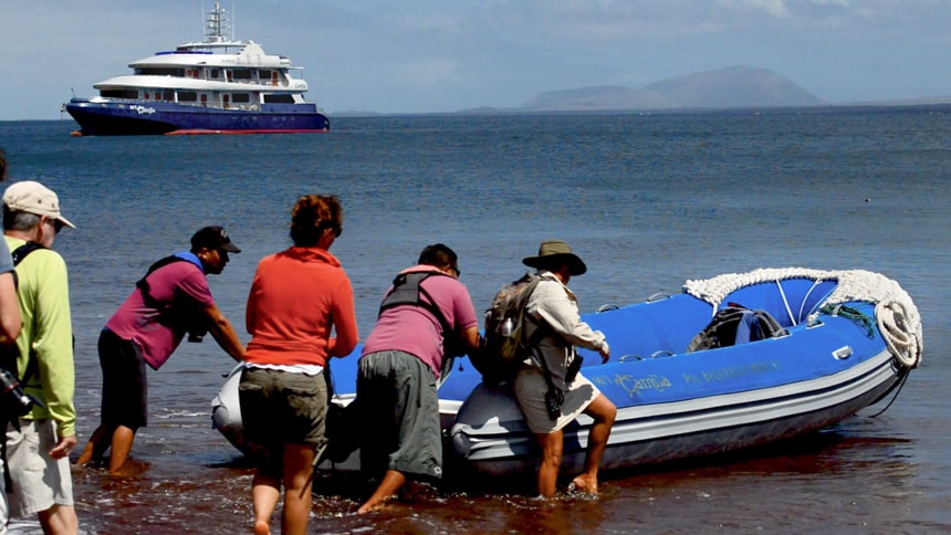 a group of travelers board an inflatable skiff or panga from the red sand beach on Rabida Island, in the distance the Camila galapagos ship floats in the ocean