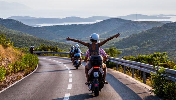 Mediterranean travelers ride scooters in Croatia on a green hillside road overlooking the mountains and ocean.