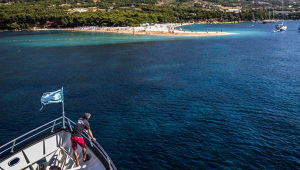 A man in red shorts looks from the bow of a small Mediterranean cruise ship with a blue flag and white deck towards a sandy beach