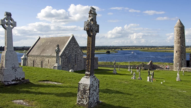 Small old stone church surrounded by green grass & stone crosses with calm river in the background on a sunny day during the Classic Ireland River Cruise.