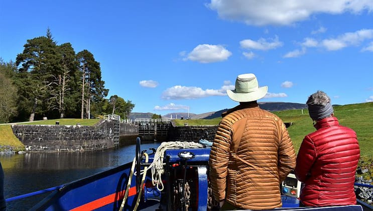 Man & woman in bright down jackets stand at the front of a hotel barge as it approaches a loch on a sunny day during the Classic Scotland Barge Cruise.