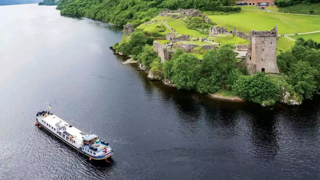 Aerial view of Urquhart Castle set along the Great Glen waterway, surrounded by bright green trees and grass with a hotel barge ship cruising by.
