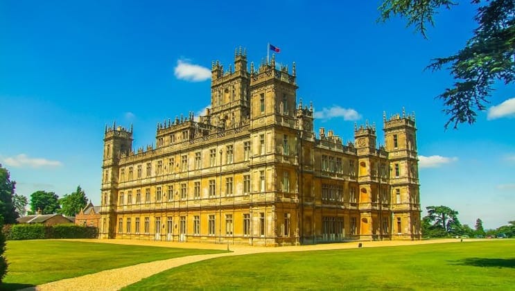 Highclere Castle with 3 stories, flags flying, green grass surrounding & blue sky, seen during a barge cruise in England.