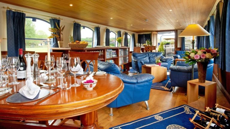 Saloon inside River Thames barge Magna Carta, with wooden table set for a meal, wooden floors, ceiling & walls, picture windows, blue leather furniture & wine rack.