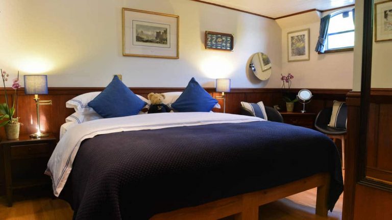 Suite with double bed, blue-&-white linens & pillows, small window, bedside tables & lamps, & wooden flooring & wainscoting aboard Magna Carta barge.