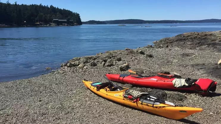 1 yellow & 1 red kayak sit on a shore of pebbles beside calm blue water with tree-lined shores in the background on a sunny day during the Voyage Through the San Juan Islands small ship cruise.
