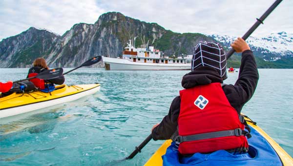 Kayakers in yellow kayaks and red life jackets paddle in Alaska towards a small wooden ship floating in front of them