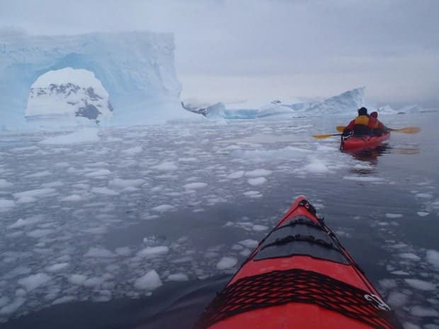 Small ship cruise guests on kayaking excursion in Antarctica close to icebergs and surrounded by ice in the water.