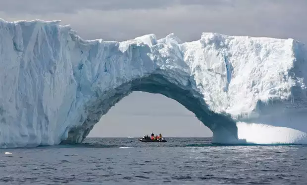 Skiff full of small cruise ship passengers underneath a large arch in the iceberg in Antarctica. 