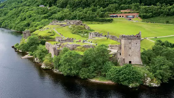 Aerial view of Urquhart Castle set along Scotland's Great Glen waterway, surrounded by bright green trees and grass.