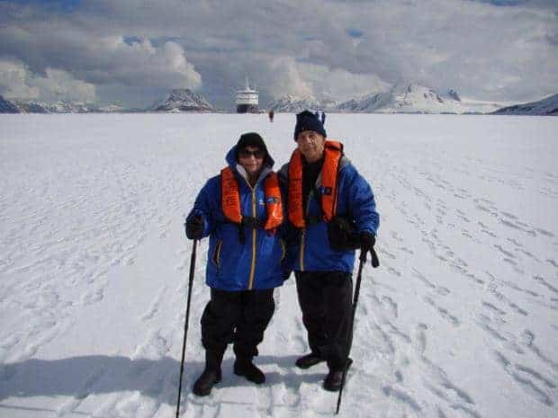 Two people on the snow in Antarctica with their walking sticks and small ship far in the background. 
