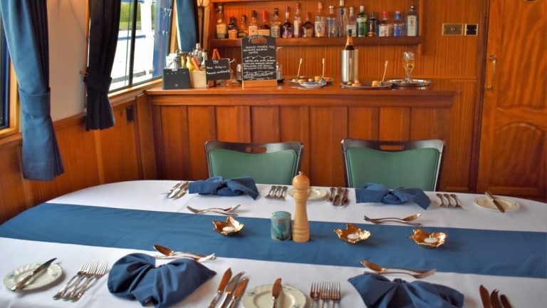 Dining room table set for a meal with white-&-blue linens & wooden bar behind aboard Scottish Highlander small riverboat.
