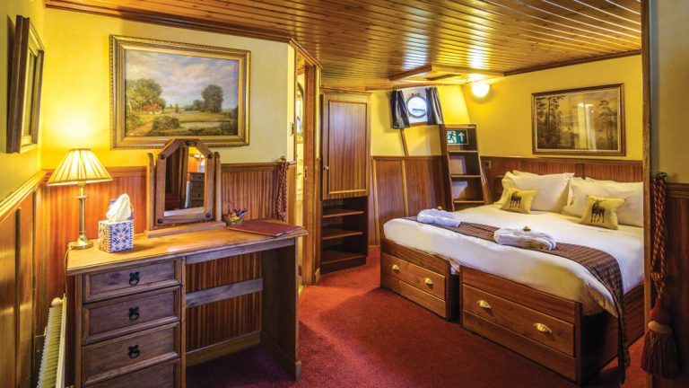 Warmly lit below-deck Cameron Suite with double bed, wooden furniture & ensuite bathroom aboard Scottish Highlander Scotland barge.