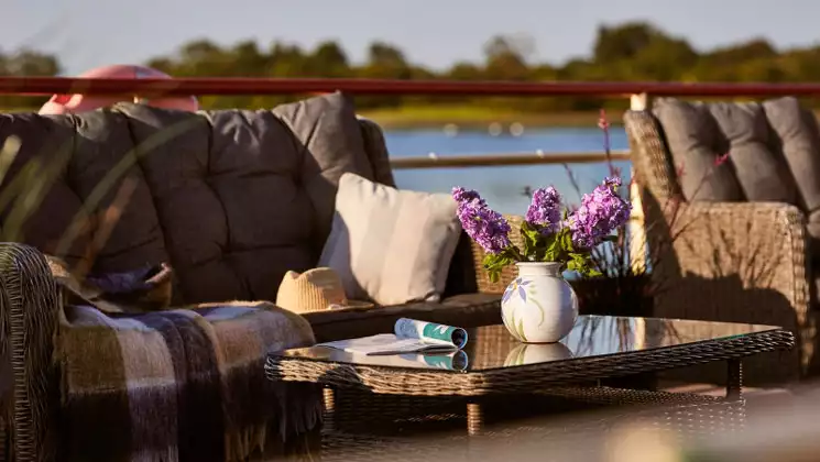 Shannon Princess barge sundeck with woven couch with cushions & coffee table with vase of lavender flowers & magazine in the sun.