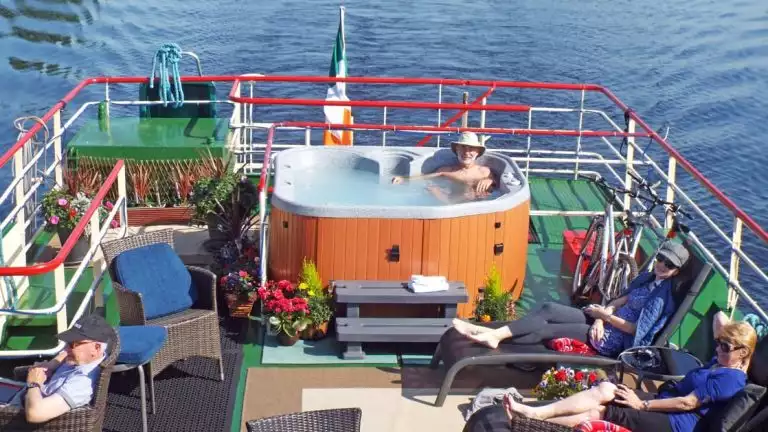 Man sits in hot tub & 3 guests sit on upper sun deck furniture while Shannon Princess barge cruises Ireland's River Shannon.