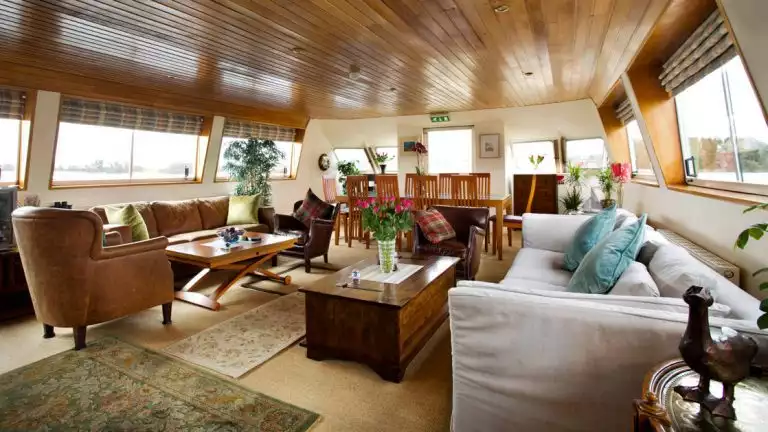 Common area with soft white & tan leather couches, wooden coffee tables, carpets, wooden ceiling, large view windows & 10-guest wooden dining table aboard Shannon Princess barge.