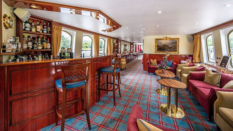 Spirit of Scotland barge saloon with checkered red & green rug, wood bar stools & bar, red & gold padded chairs & plenty of light.