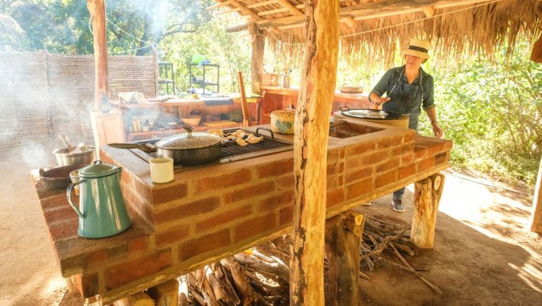 Woman stands over a brick stove making tortillas under a thatched roof on a sunny day at Camp Cecil de la Sierra.