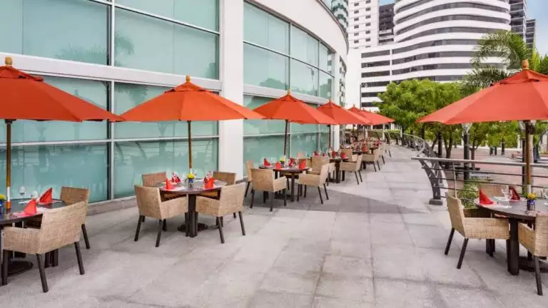 Exterior of Rio Grande Restaurant at Wyndham Guayaquil Hotel, with red-orange umbrellas & wicker chairs.