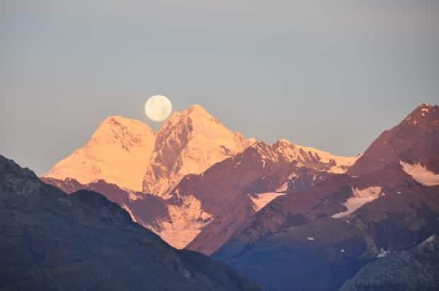 Full moon sitting atop the peak of a mountain range at dusk.