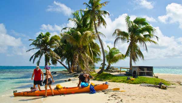 3 kayakers unload an orange kayak on a sandy beach under palm trees & blue sky during a Belize cruise excursion.