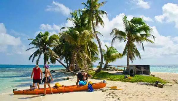 3 kayakers unload an orange kayak on a sandy beach under palm trees & blue sky during a Belize cruise excursion.