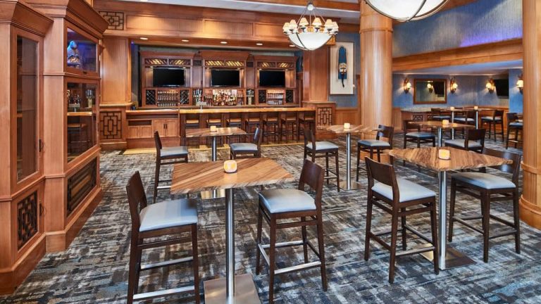 The bar and lounge of the Hilton Downtown Anchorage. Hit top tables anc chairs are set around the room with a wooden bar with flat screen tvs.