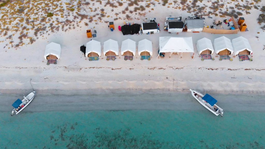 A oceanfront row of Luxury glamping tent at Camp Cecil on Isla Espritu Santo in Baja, An aerial photo taken from above showing teal ocean with two boats floating at the shoreline of a white sand beach.