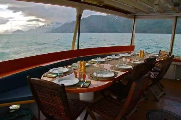 Dinner table aboard the small ship Sea Wolf surrounded by windows looking at the mountains of Alaska. 