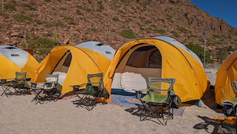 Yellow & white, REI-brand dome tents set up on a beach with green camp chairs & blue mats in front, for tent camping Baja.