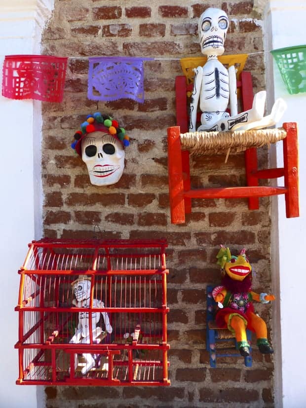 Mexican Day of the Dead masks and skeletons sitting on a chair and in a bird cage on a brick wall with colorful flags.