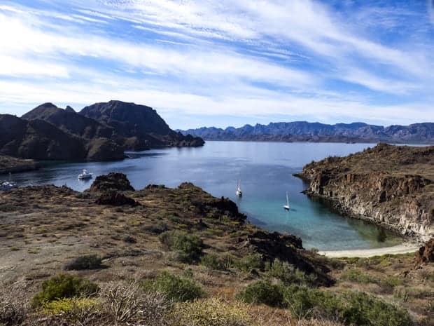 A view of from a top a rocky cliff overlooking a sandy beach and mountain ranges with 2 sailboats and small ship cruise anchored.