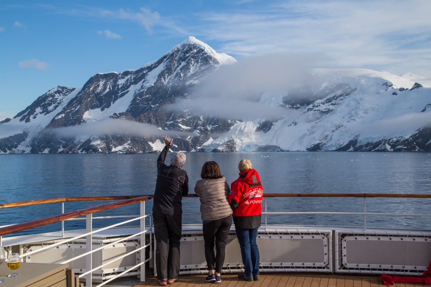 Three guests stand at the front observation deck of a luxury small ship staring at the icy mountain landscape of Antarctifca