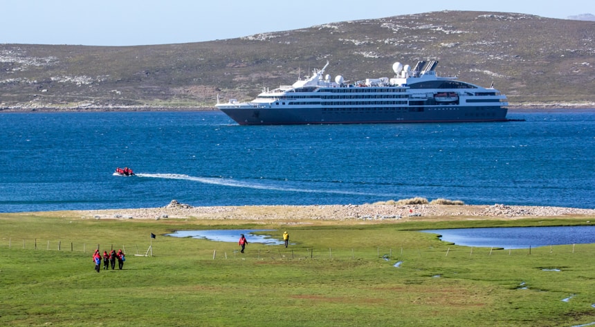 L'Austral, a luxury ship is floating in the blue ocean water as a group of people in red parkas walk towards the shore from a bright green grassy meadow