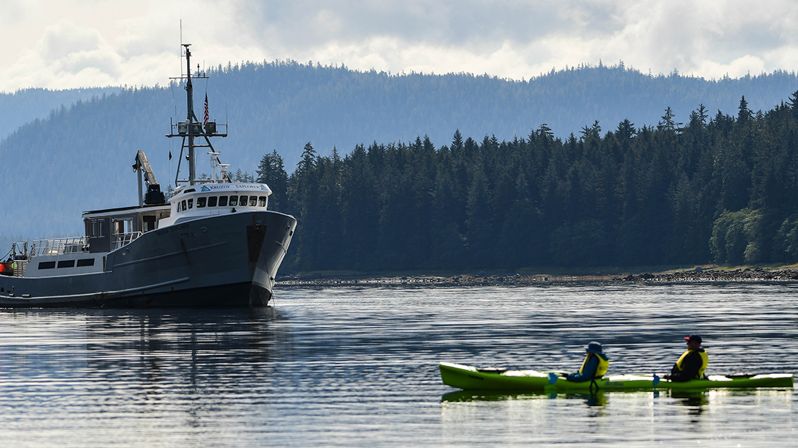 Starboard exterior of the Kruzof Explorer Alaska small ship, anchored on a cloudy day with a single kayak in the water in front of it