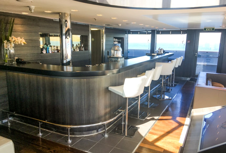 The bar area aboard L'Austral and her fleet of luxury ponant sister ships. grey curved counter top with white leather bar seats with silver accents are surrounded by glass windows to an outside viewing area.