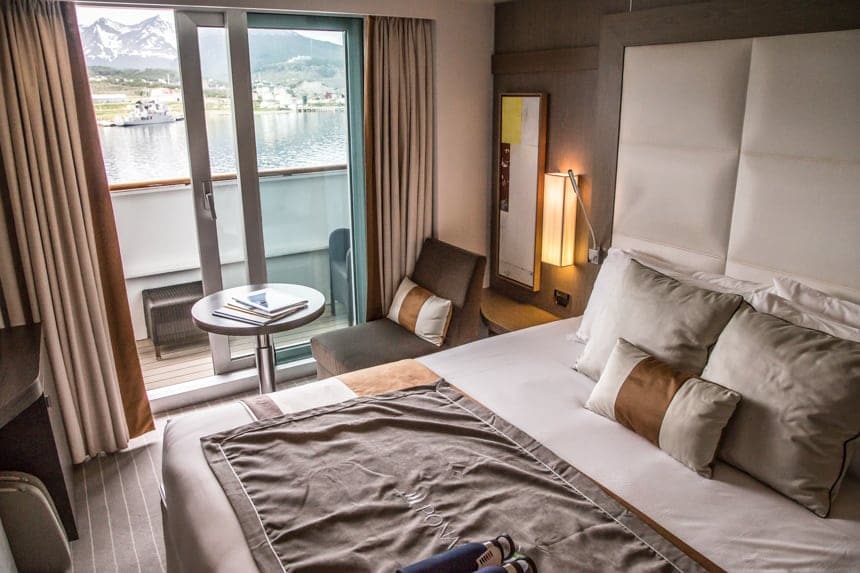 A cabin inside L'Austral and her fleet of luxury ponant sister ships. Neutral color pallet of white grey and golds are used throughout. A white leather tall headboard sits above the king bed, next to a small accent chair and coffee table. A sliding glass door leads to a private balcony