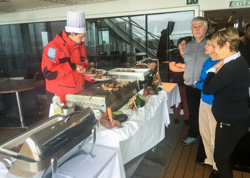 Aboard luxury small ship L'Austral a chef wearing a red parka and tall chefs hat stands behind an outdoor BBQ table and serves food to guests standing in line